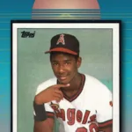 gary_pettis_80s_title_v4 » Cards Mania 1985 Topps baseball card #497 showing a young player in a California Angels uniform, pointing toward the camera, with the card caption "Angels – Gary Pettis" at the bottom. The card is set against a retro 1980s synthwave background with a teal-to-coral gradient sky and a sliced sun. Large overlaid text above the card reads "He is not Gary Pettis!" — a nod to the famous printing error in which the pictured player is widely believed to be Gary's younger brother rather than Gary himself.